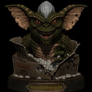 Puede incluir: Una criatura verde y escamosa con orejas grandes, ojos naranjas y un mech&oacute;n de pelo blanco que emerge de una caja de cart&oacute;n. La caja presenta el texto FRAGILE y HANDLE WITH CARE. Una placa en la base muestra el texto Nostalgiahorror3d en oro.