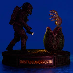 Op de afbeelding: Een donker, gedetailleerd beeldje van een astronaut in ruimtepak met een zaklamp, staand voor een open, getextureerd ei waaruit een wezen tevoorschijn komt. De basis toont de tekst NOSTALGIAHORROR3D in gouden letters.