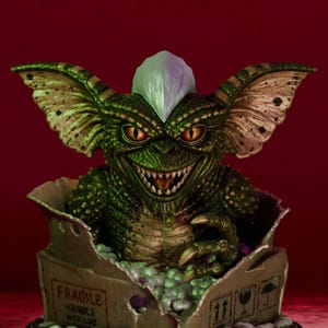 Puede incluir: Una criatura verde texturizada con orejas grandes, dientes afilados y una cresta blanca que emerge de una caja de cart&oacute;n con la etiqueta FRAGILE. La caja se asienta sobre una base circular negra con una placa met&aacute;lica que dice Nostalgiahorror3d.