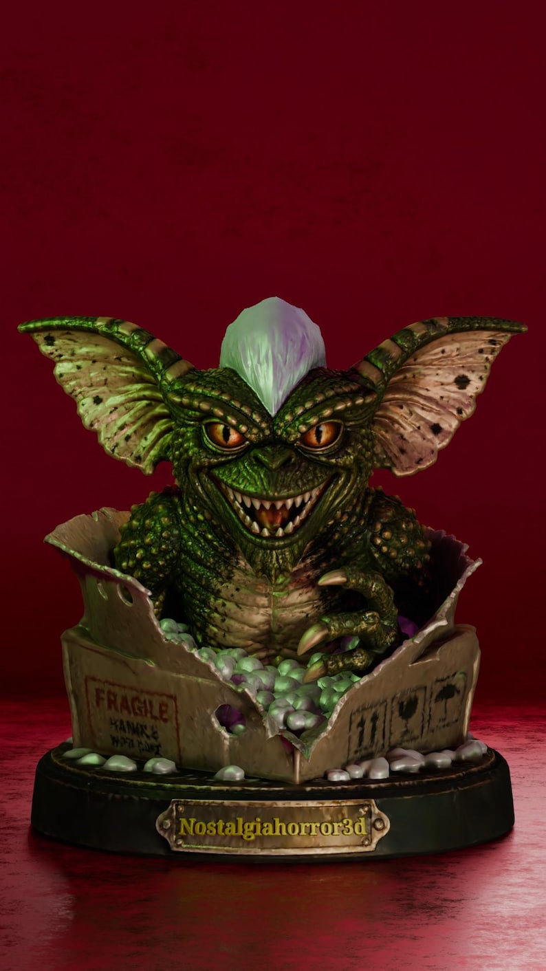 Puede incluir: Una criatura verde texturizada con orejas grandes, dientes afilados y una cresta blanca que emerge de una caja de cart&oacute;n con la etiqueta FRAGILE. La caja se asienta sobre una base circular negra con una placa met&aacute;lica que dice Nostalgiahorror3d.