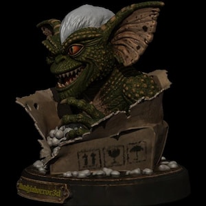 Puede incluir: Una criatura verde y escamosa con orejas grandes, dientes afilados y pelo blanco que emerge de una caja de cart&oacute;n rota. La caja tiene tres iconos de env&iacute;o negros y se asienta sobre una base circular con una placa dorada NightHorror3d.