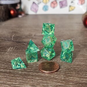 May include: A collection of translucent green dice with blue numbering, arranged on a wooden surface. The dice are various shapes, including a pyramid, a cube, and a multi-sided die. A penny is next to the dice.