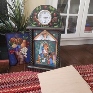 May include: A decorative clock with a painted nativity scene and a round clock face. The clock is adorned with painted apples and leaves. A painting of a pumpkin and other fruits is to the left. A wooden board is in the foreground.