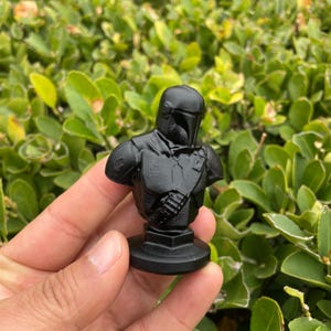 Op de afbeelding: Zwarte buste van een Mandalorian-personage, met helm en pantserdetails. Het beeldje staat op een ronde basis en wordt in een hand gehouden, tegen een achtergrond van groen gebladerte. De buste is effen matzwart.