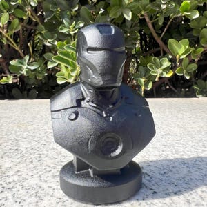 Puede incluir: Busto negro de un superh&eacute;roe, con cabeza con casco y armadura pectoral. La escultura es de material texturizado y se apoya sobre una base redonda. El busto est&aacute; frente a follaje verde.