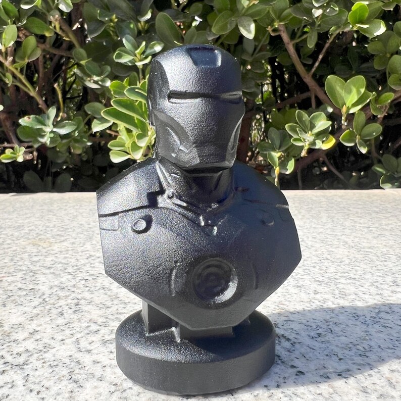 Puede incluir: Busto negro de un superh&eacute;roe, con cabeza con casco y armadura pectoral. La escultura es de material texturizado y se apoya sobre una base redonda. El busto est&aacute; frente a follaje verde.