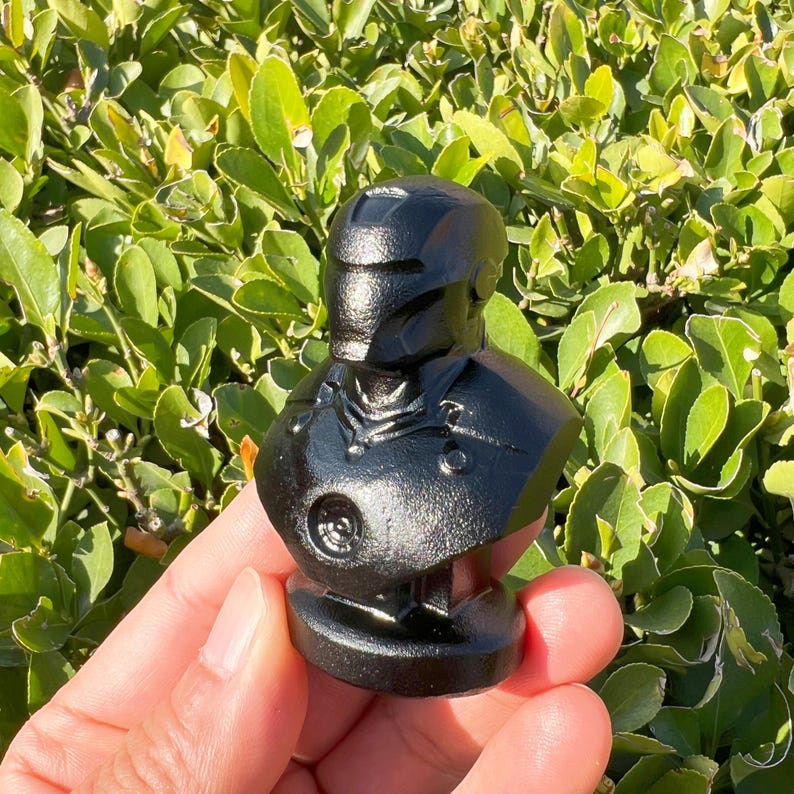 May include: A black bust of a superhero with a helmet and armor. The bust is made of a shiny, dark material and has detailed features. The bust is a collectible item.
