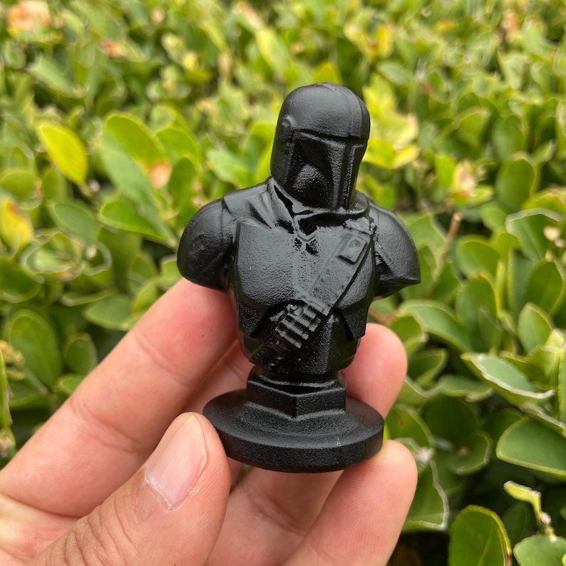 Op de afbeelding: Een zwarte buste van een personage uit een sciencefictionreeks. De figuur draagt een helm en harnas, met een gedetailleerd borststuk en een riem over de torso. De buste is gemonteerd op een cirkelvormige basis.