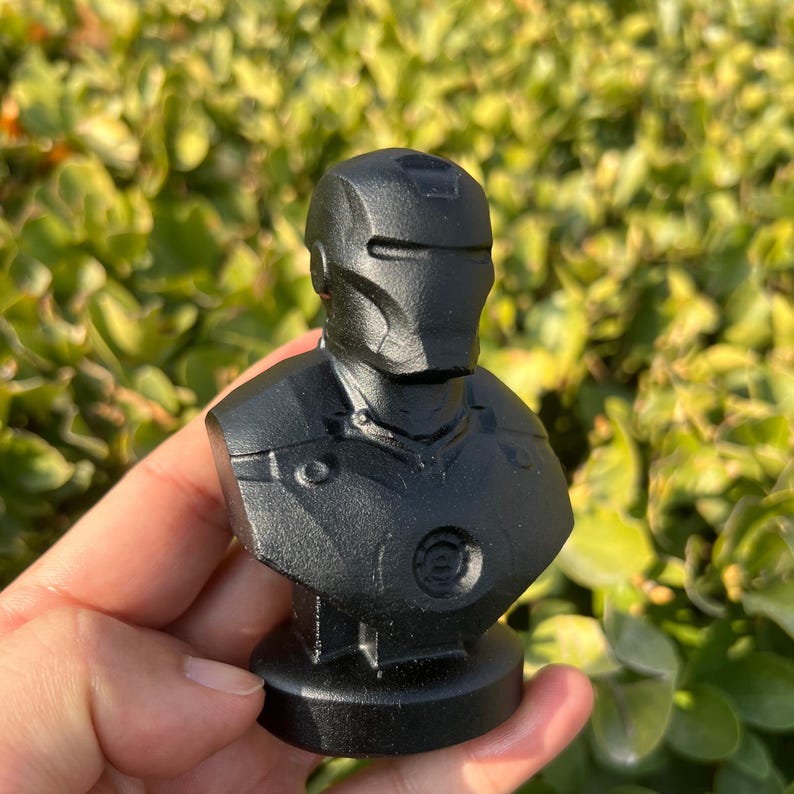 May include: A black bust of Iron Man, featuring the character's helmet and armor details. The sculpture is mounted on a circular base. The bust is held in a hand against a backdrop of green foliage.