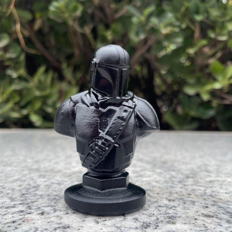Op de afbeelding: Een zwarte buste van een Mandalorian-personage, met helm en pantserdetails. De sculptuur is gemonteerd op een zwarte basis. Het is een verzamelobject, waarschijnlijk een fan-creatie.