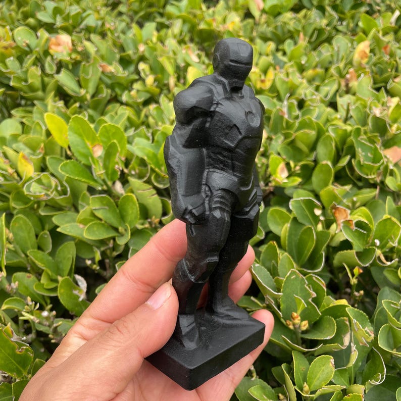 May include: A black Iron Man figurine, standing on a rectangular base. The figure is detailed with armor plating and a helmet. The statue is held in front of a backdrop of green foliage.