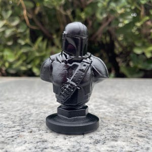 Op de afbeelding: Een zwarte Mandalorian buste beeldje. Het beeldje toont de helm en het pantser van het personage, met een gedetailleerde schouderriem en borstplaat. De buste is gemonteerd op een cirkelvormige basis en het algehele ontwerp is strak en modern.