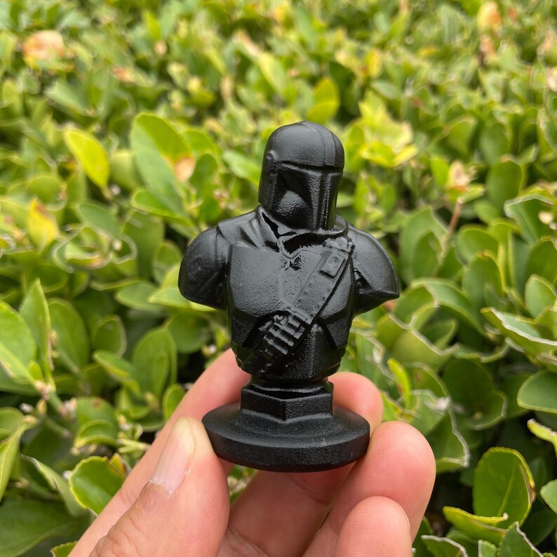 Op de afbeelding: Een zwarte Mandalorian buste. Het beeldje toont in detail het pantser en de helm van het personage, gemonteerd op een kleine, vierkante basis. De buste is gemaakt van een solide, matzwart materiaal.