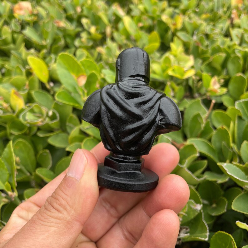 Op de afbeelding: Een kleine, zwarte buste-sculptuur van een figuur met helm en cape. De sculptuur wordt in een hand gehouden tegen een achtergrond van groen gebladerte. De buste is op een cirkelvormige basis gemonteerd.