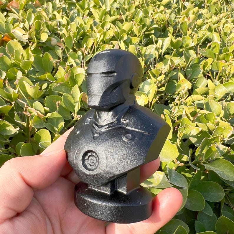 May include: A black bust of a superhero, featuring a detailed helmet and armor design. The sculpture is held in a hand against a backdrop of green foliage. The bust is a collectible item.