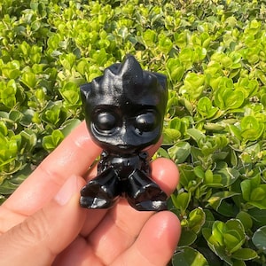 May include: A small, black figurine of a character with large eyes and a distinctive head shape. The figurine is held in a hand against a backdrop of green foliage. The character's features are clearly defined, and the overall appearance is smooth and glossy.
