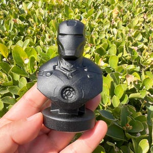 Puede incluir: Un busto negro de Iron Man, que representa el casco y la armadura del pecho del personaje. El busto est&aacute; sobre una base circular y se sostiene en una mano. El fondo es una densa disposici&oacute;n de hojas verdes.