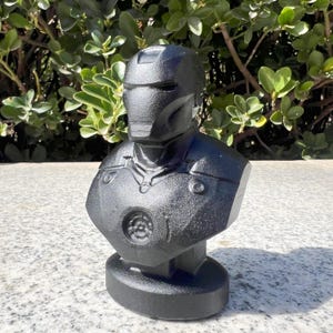 Puede incluir: Busto negro de un superh&eacute;roe con dise&ntilde;o de casco y armadura. La escultura est&aacute; hecha de un material negro mate y se asienta sobre una base redonda. Representaci&oacute;n detallada de un personaje de c&oacute;mic.
