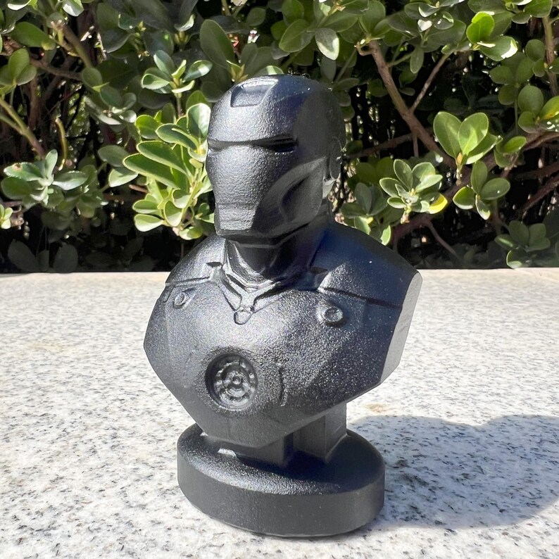 Puede incluir: Busto negro de un superh&eacute;roe con dise&ntilde;o de casco y armadura. La escultura est&aacute; hecha de un material negro mate y se asienta sobre una base redonda. Representaci&oacute;n detallada de un personaje de c&oacute;mic.