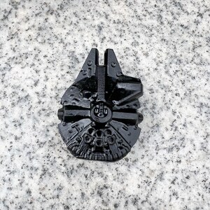 Puede incluir: Un modelo negro y detallado de la nave espacial Halcón Milenario. El modelo está hecho de un material oscuro y presenta intrincados detalles de la superficie, incluyendo paneles, torretas y otros elementos mecánicos. La forma icónica de la nave es claramente visible.