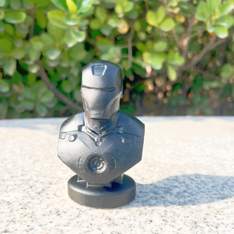 May include: A black bust of Iron Man, featuring the character's helmet and chest armor. The sculpture is set on a circular base and is made of a matte, dark material. The background is blurred, with green foliage and a light-colored surface.