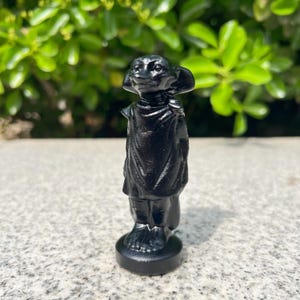 Puede incluir: Una estatuilla negra de una criatura con orejas grandes y una prenda drapeada. La estatua está sobre una base redonda. El fondo es follaje verde borroso.
