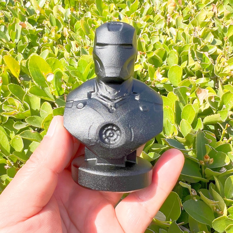 Puede incluir: Busto negro de Iron Man, con un casco y una placa pectoral detallados. La escultura se encuentra sobre un fondo de follaje verde, destacando el dise&ntilde;o ic&oacute;nico del superh&eacute;roe. El busto mide unos 10 cm de altura.