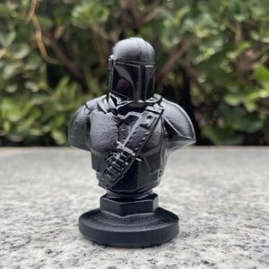 Op de afbeelding: Een zwarte buste van een Mandalorian-personage, met een helm en harnas, gemonteerd op een ronde basis. De figuur heeft een gedetailleerd ontwerp, inclusief een schouderriem. De buste is gemaakt van glanzend materiaal.