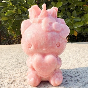 Könnte beinhalten: Eine rosa geschnitzte Figur einer Zeichentrickkatze, die ein Herz hält. Die Katze hat eine Schleife auf dem Kopf und besteht aus einem glatten, polierten Material. Die Figur steht aufrecht und hat ein detailliertes Design.
