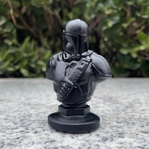 Op de afbeelding: Zwarte buste van een Mandalorian krijger, met helm, schouderpantser en bandelier. De sculptuur staat op een ronde basis. De achtergrond is wazig groen gebladerte.