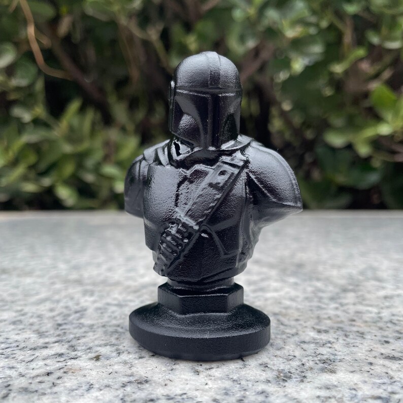 Op de afbeelding: Zwarte buste van een Mandalorian krijger, met helm, schouderpantser en bandelier. De sculptuur staat op een ronde basis. De achtergrond is wazig groen gebladerte.