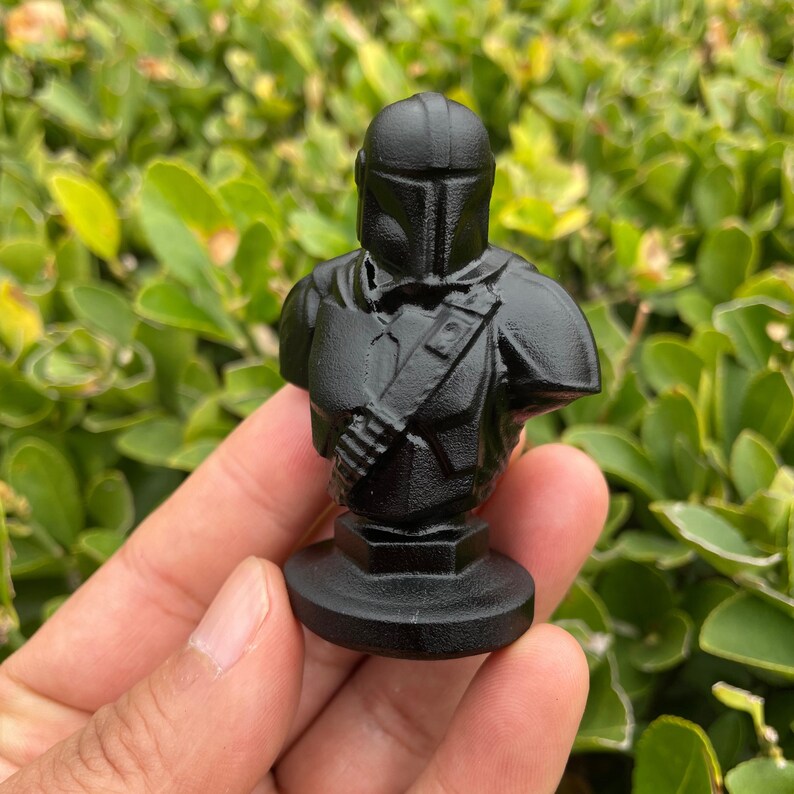 Op de afbeelding: Een zwarte buste van een Mandalorian-personage, met een helm en pantserdetails. De figuur is gemonteerd op een cirkelvormige basis. De buste wordt in een hand gehouden tegen een achtergrond van groen gebladerte.
