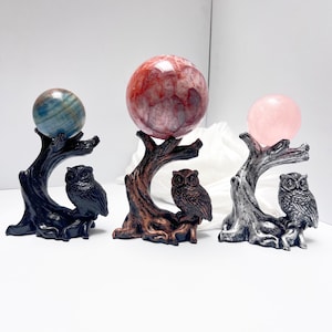 May include: Three decorative stands shaped like tree branches with perched owls, each holding a polished stone sphere. The left stand is black with a blue sphere, the centre is copper with a red sphere, and the right is silver with a pink sphere.