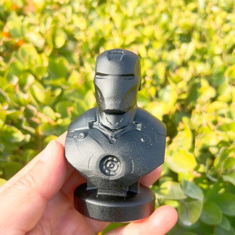 May include: A black bust of Iron Man, featuring the character's helmet and chest armor. The sculpture is made of a solid, matte black material and is mounted on a circular base. The bust is a collectible item.
