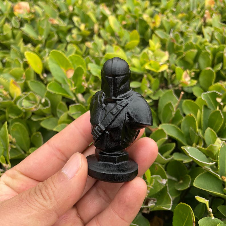 Op de afbeelding: Een zwarte Mandalorian buste beeldje. Het beeldje is een gedetailleerde weergave van het pantser en de helm van het personage. De buste is gemonteerd op een ronde basis. De achtergrond is wazig groen gebladerte.