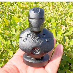 Puede incluir: Un busto negro de Iron Man. La estatua presenta una representaci&oacute;n detallada del casco y la armadura del pecho del personaje. El busto se sostiene en una mano sobre un fondo de follaje verde.