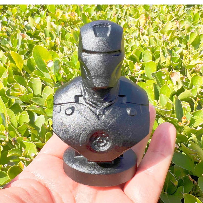 Puede incluir: Un busto negro de Iron Man. La estatua presenta una representaci&oacute;n detallada del casco y la armadura del pecho del personaje. El busto se sostiene en una mano sobre un fondo de follaje verde.