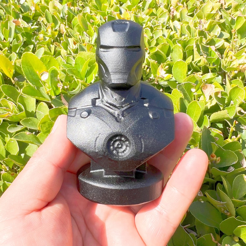 Puede incluir: Busto negro de Iron Man, con el casco y la armadura del pecho del personaje. La escultura se sostiene en una mano sobre un fondo de follaje verde. El busto tiene un acabado mate y est&aacute; montado sobre una peque&ntilde;a base.