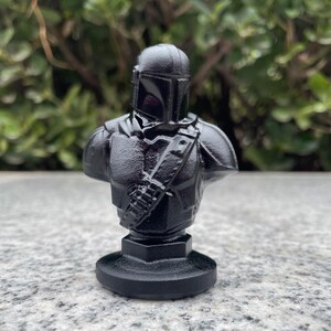 Op de afbeelding: Zwarte buste van een Mandalorian krijger, met helm, harnas en een schouderriem met een tasje. De sculptuur staat op een ronde basis. De buste is effen zwart en heeft een getextureerd oppervlak.