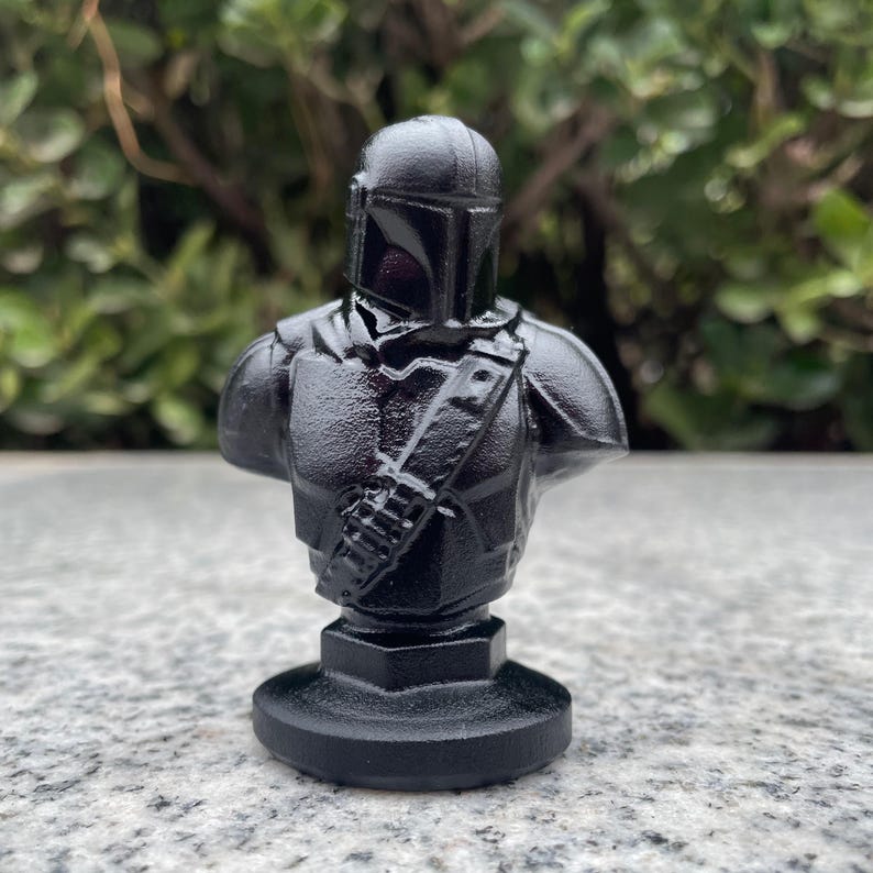 Op de afbeelding: Zwarte buste van een Mandalorian krijger, met helm, harnas en een schouderriem met een tasje. De sculptuur staat op een ronde basis. De buste is effen zwart en heeft een getextureerd oppervlak.