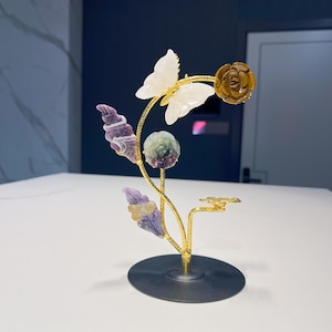 May include: A decorative floral sculpture with a butterfly, a rose, and leaves. The butterfly has white wings, and the rose is brown. The leaves are purple and yellow. The sculpture is mounted on a gold stem and a black circular base.