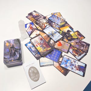 May include: A collection of tarot cards with colorful illustrations, scattered on a white surface. A metal tin and a guidebook, both labeled "Witches Tarot", are also present. The tin features a woman holding a torch. A clear, cylindrical crystal is also visible.