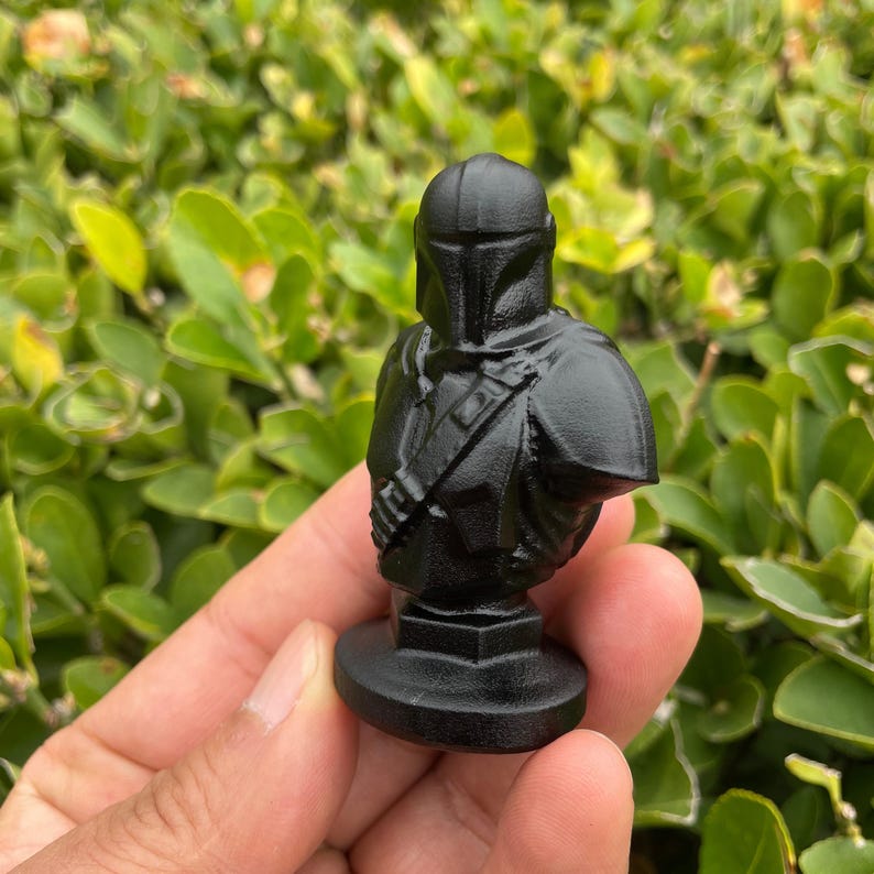 Op de afbeelding: Een zwarte Mandalorian buste. Het beeldje toont in detail de helm en het pantser van het personage, gemonteerd op een vierkante basis. De buste staat tegen een achtergrond van groen gebladerte.