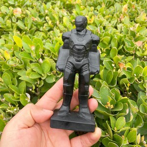 May include: A black figurine of a superhero in a standing pose. The figurine has a detailed design, including a helmet, chest plate, and leg armor. The figure is held in a hand against a backdrop of green foliage.
