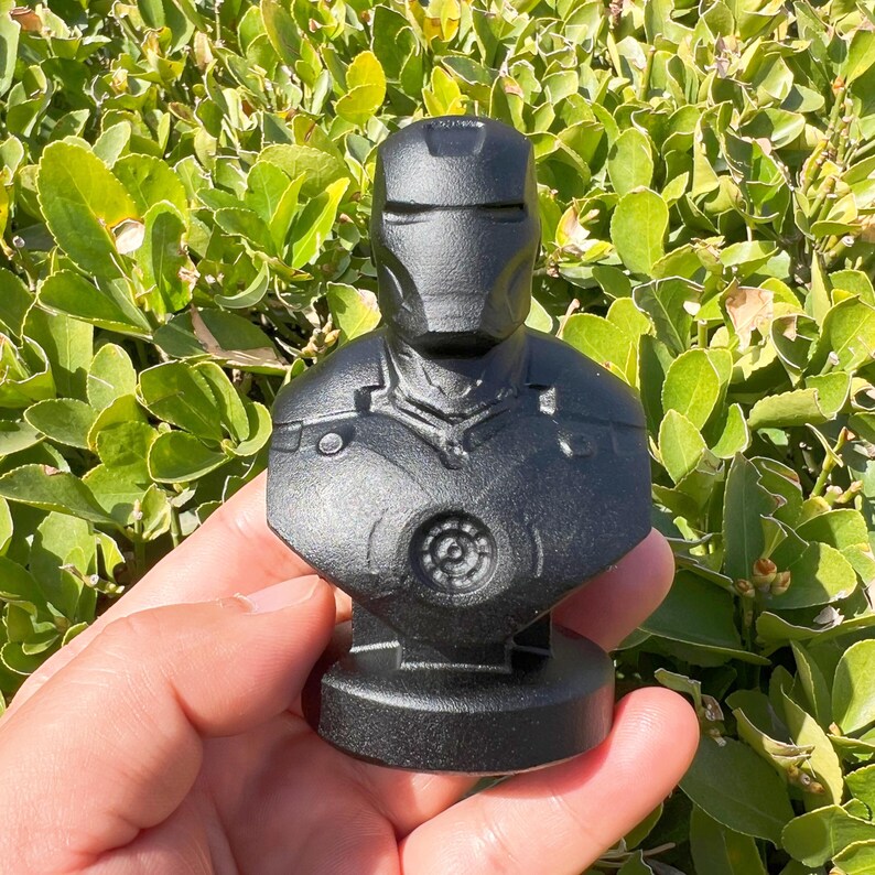 Puede incluir: Un busto negro de Iron Man, que representa el casco y la armadura del pecho del personaje. La escultura se sostiene en una mano, sobre un fondo de follaje verde. El busto tiene un acabado mate y un dise&ntilde;o detallado.
