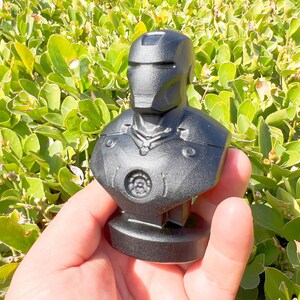 Puede incluir: Un busto negro de Iron Man, que presenta el casco y la armadura del pecho del personaje. La escultura se presenta sobre un fondo de follaje verde, destacando el dise&ntilde;o detallado y la textura del traje del superh&eacute;roe.