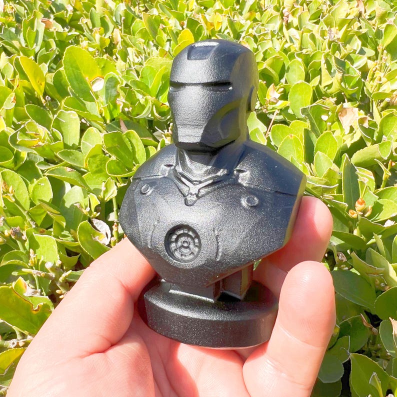 Puede incluir: Un busto negro de Iron Man, que presenta el casco y la armadura del pecho del personaje. La escultura se presenta sobre un fondo de follaje verde, destacando el dise&ntilde;o detallado y la textura del traje del superh&eacute;roe.
