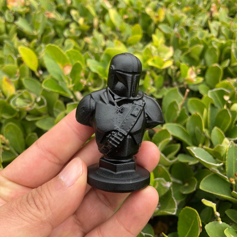 Op de afbeelding: Een zwarte buste van een Mandalorian-personage, met helm en pantserdetails. Het beeldje wordt tegen een achtergrond van groen gebladerte gehouden, wat het gedetailleerde ontwerp en de textuur van de sculptuur laat zien.