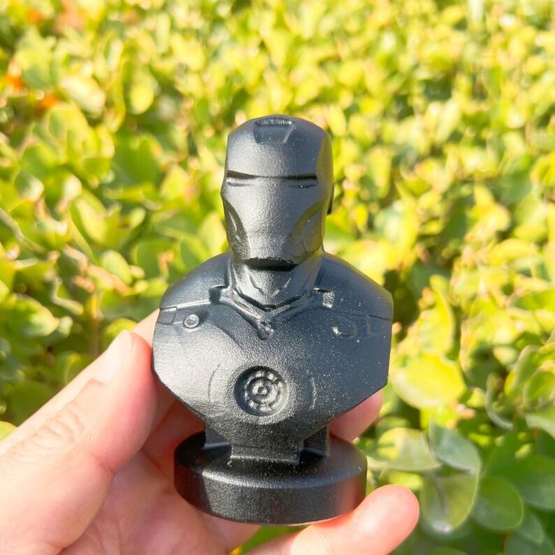 May include: A black bust of Iron Man, featuring the character's helmet and chest armor. The sculpture is made of a solid, matte black material and is mounted on a circular base. The bust is a collectible item.