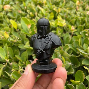 Op de afbeelding: Zwarte buste van een Mandalorian-personage, met helm en harnas. De figuur is gemonteerd op een ronde basis. De buste wordt in een hand gehouden tegen een achtergrond van groen gebladerte.